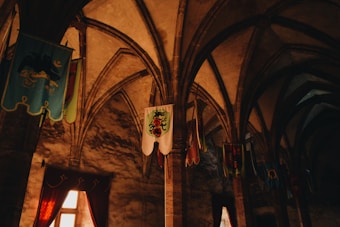A dimly lit medieval hall with arched ceilings and stone columns. The walls are adorned with colorful banners featuring various heraldic symbols. Warm light streams in through small windows, casting shadows and adding to the ancient ambiance.