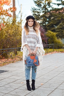 A young woman stands outdoors, wearing a patterned poncho, distressed jeans, and a black hat. She holds a blue bag with colorful patches, set against a backdrop of autumn foliage.