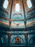 An ornate interior with intricate architectural details, featuring classical sculptures and reliefs on the walls. The ceiling rises high with large, oval windows allowing light to create a dramatic ambiance. The lavish decor includes gold accents and elaborate carvings.