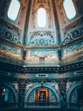 An ornate interior with intricate architectural details, featuring classical sculptures and reliefs on the walls. The ceiling rises high with large, oval windows allowing light to create a dramatic ambiance. The lavish decor includes gold accents and elaborate carvings.