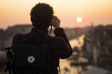 A person with curly hair is photographed from behind, holding a camera and taking a picture of a scenic sunset over a cityscape. The silhouette of the person is highlighted against the warm colors of the sunset, with a large backpack also visible, suggesting they are a traveler or photographer.