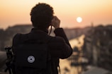 A person with curly hair is photographed from behind, holding a camera and taking a picture of a scenic sunset over a cityscape. The silhouette of the person is highlighted against the warm colors of the sunset, with a large backpack also visible, suggesting they are a traveler or photographer.