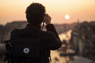 A person with curly hair is photographed from behind, holding a camera and taking a picture of a scenic sunset over a cityscape. The silhouette of the person is highlighted against the warm colors of the sunset, with a large backpack also visible, suggesting they are a traveler or photographer.