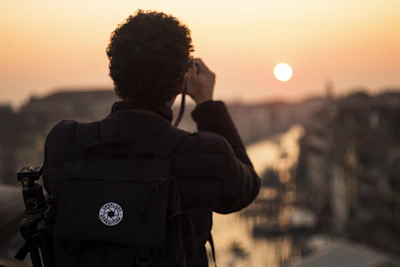 A person with curly hair is photographed from behind, holding a camera and taking a picture of a scenic sunset over a cityscape. The silhouette of the person is highlighted against the warm colors of the sunset, with a large backpack also visible, suggesting they are a traveler or photographer.