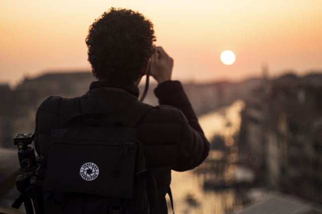 A person with curly hair is photographed from behind, holding a camera and taking a picture of a scenic sunset over a cityscape. The silhouette of the person is highlighted against the warm colors of the sunset, with a large backpack also visible, suggesting they are a traveler or photographer.