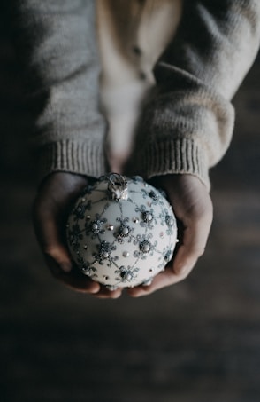 A pair of hands cradles an ornate, decorative ornament, which is spherical and adorned with intricate silver patterns and small pearl-like accents. The person is wearing a cozy, knit sweater, and the background is softly blurred, creating a warm and intimate atmosphere.