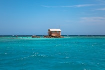 A small, secluded wooden house stands on a rocky islet surrounded by clear turquoise water, with a bright blue sky above. The house has a white roof and an antenna. The surrounding water is calm and inviting, indicating a peaceful, remote location.