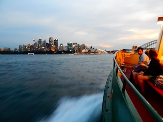 A ferry is traveling across a body of water towards a cityscape featuring illuminated skyscrapers and an iconic sail-shaped building. Several people are seated on the ferry, enjoying the view, with some engaged in conversation. The sky is a mix of blue and orange hues, suggesting either dawn or dusk.