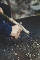 A person using a knife to carve or whittle a piece of wood, with wood shavings scattered on a dark surface. The person's hands are seen holding the piece of wood firmly. The background is blurred, focusing on the hands and the carving activity.