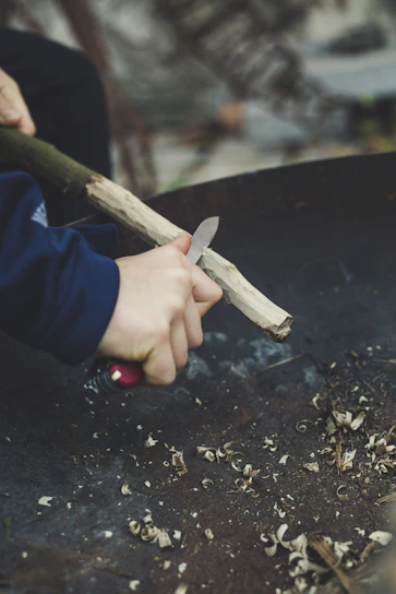 Close-up of a hand carving a wooden spoon, surrounded by wood shavings and natural light.