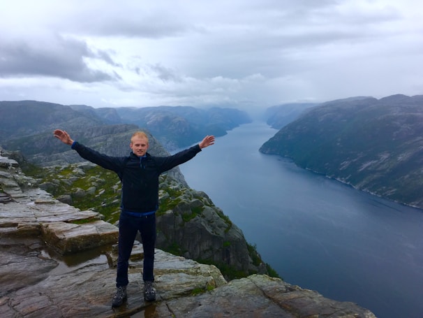 A person stands on a rocky cliff with arms outstretched, overlooking a expansive, winding fjord surrounded by steep cliffs. The sky is overcast, adding a dramatic atmosphere to the scene.