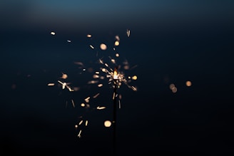 A sparkler lit in the dark, with glowing sparks radiating outward. The background is mostly black, highlighting the bright sparks and fiery particles.