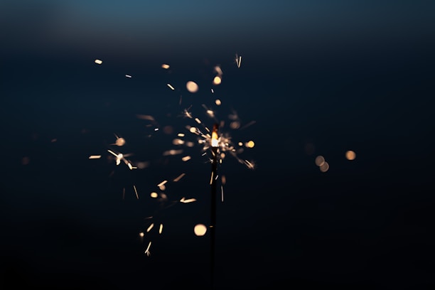 A sparkler lit in the dark, with glowing sparks radiating outward. The background is mostly black, highlighting the bright sparks and fiery particles.