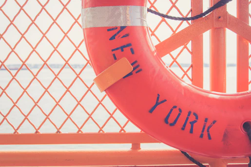 An orange life preserver with the words 'New York' printed on it is mounted on an orange metal grid. The background features a body of water with a faint view of a bridge or coastline.