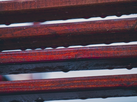 Lines of horizontal wooden planks with a reddish hue and visible texture, possibly wet or coated with a glossy finish. The planks are spaced slightly apart, allowing glimpses of a lighter background.