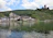 A picturesque village nestled by a calm river, with quaint buildings featuring traditional architecture. In the background, lush green hills rise, partially covered by vineyards. A historic castle ruin is perched atop the hill, adding an element of medieval charm. The sky is partly cloudy, creating a serene and tranquil atmosphere.
