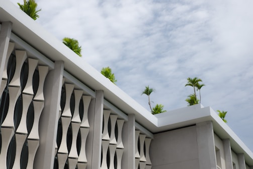 A modern building facade featuring stylized, geometric concrete panels. The roof is adorned with several small palm trees that add a touch of greenery against a backdrop of partially cloudy skies.