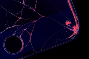 A close-up of a shattered screen on a dark surface, with cracks radiating from a central point and edges illuminated in a neon-like glow.