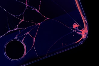 A close-up of a shattered screen on a dark surface, with cracks radiating from a central point and edges illuminated in a neon-like glow.