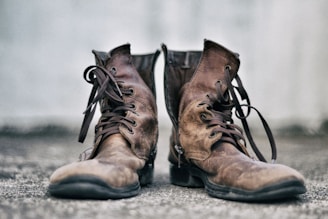 A pair of well-worn brown leather boots with dark laces sits on a textured surface. The background is a soft, blurred gray, highlighting the rustic and rugged nature of the boots.