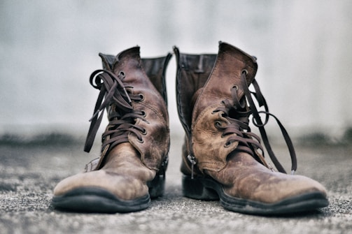 A pair of well-worn brown leather boots with dark laces sits on a textured surface. The background is a soft, blurred gray, highlighting the rustic and rugged nature of the boots.