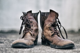 A pair of well-worn brown leather boots with dark laces sits on a textured surface. The background is a soft, blurred gray, highlighting the rustic and rugged nature of the boots.