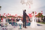 A couple in wedding attire stands by a pool holding hands, surrounded by a crowd of people releasing red balloons into the sky. The scene is festive with a backdrop of greenery and a partly cloudy sky.