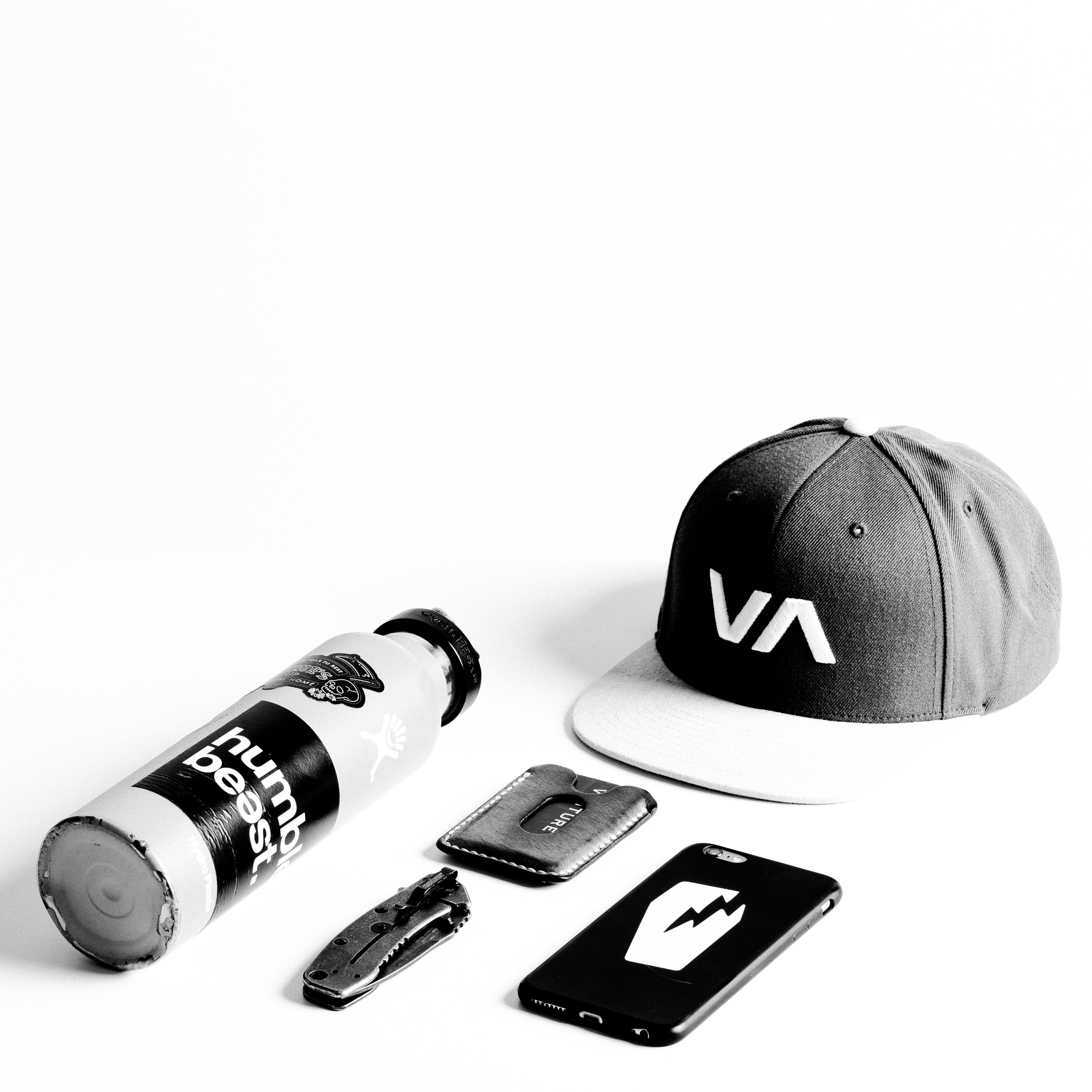 Black and white photograph of a hat, a cellphone, a money clip, a pocket knife and a water bottle.