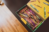 An open box of Crayola crayons filled with an assortment of vibrant colors. The box is resting on a dark surface, and several crayon sticks are scattered on the lid which features a yellow polka dot pattern. The hardwood floor in the background adds a warm tone to the image.