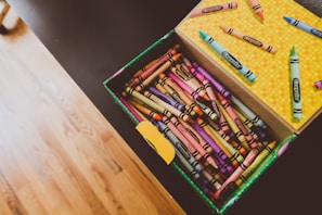 An open box of Crayola crayons filled with an assortment of vibrant colors. The box is resting on a dark surface, and several crayon sticks are scattered on the lid which features a yellow polka dot pattern. The hardwood floor in the background adds a warm tone to the image.