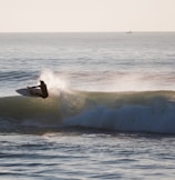 A surfer is skillfully riding a wave, with the ocean stretching out into the distance under a clear sky. The movement of the board against the water creates splashes, capturing the energy and thrill of surfing.