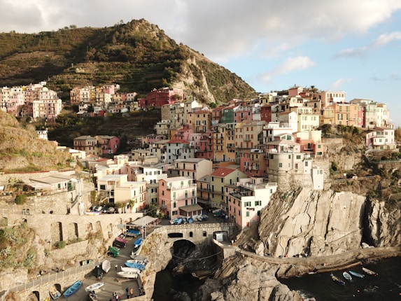 A picturesque coastal village with colorful houses built on steep cliffs overlooking the sea. The houses are tightly packed and display a variety of pastel colors, contrasting with the rugged natural landscape. The area is bathed in warm sunlight, creating a serene atmosphere.