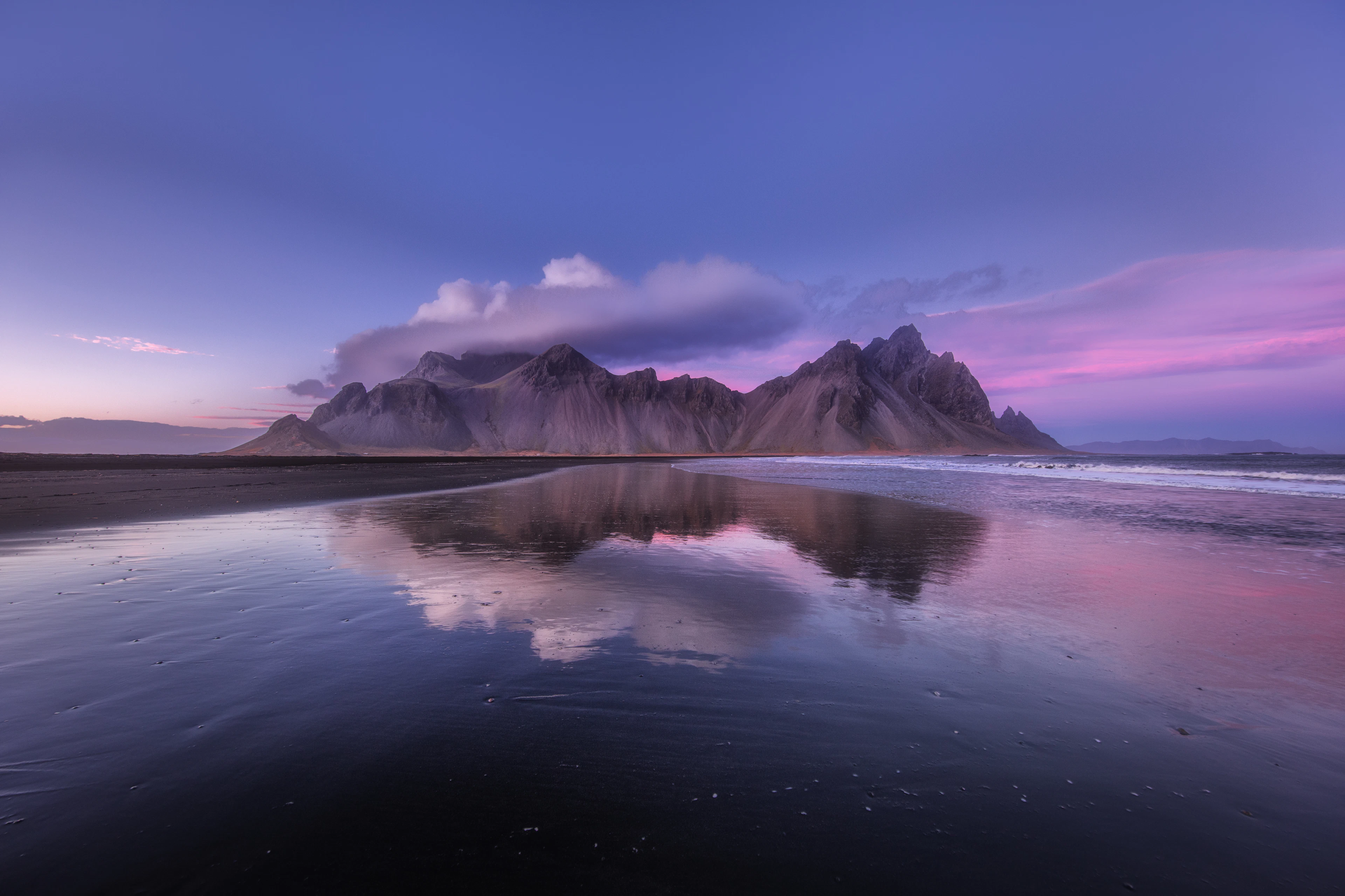 Jagged Vestrahorn mountain glows pink at dusk, its peak mirrored in wet black sand.