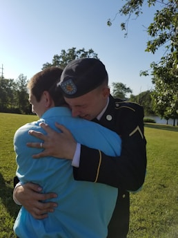 Two people embrace warmly in a grassy outdoor setting. One is wearing a military uniform, complete with a beret, and the other is dressed in a light blue shirt. Trees and a pond can be seen in the background, with a clear blue sky overhead.