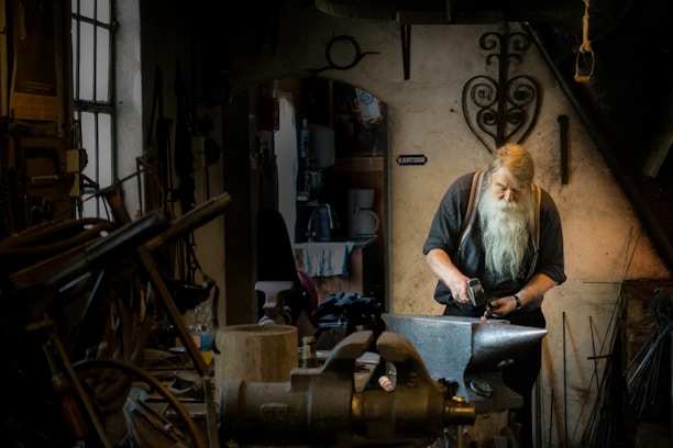 A bearded person working intently at a forge in a dimly lit workshop. The environment is filled with various tools and metalworking equipment. The scene conveys a sense of craftsmanship and dedication.