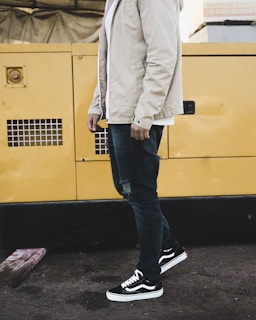 A person is standing next to a large yellow metal structure, possibly a generator. They are wearing a light beige jacket, blue jeans with distressing, and black sneakers with white soles. The ground is dark concrete, and there is a piece of wood on the ground nearby.