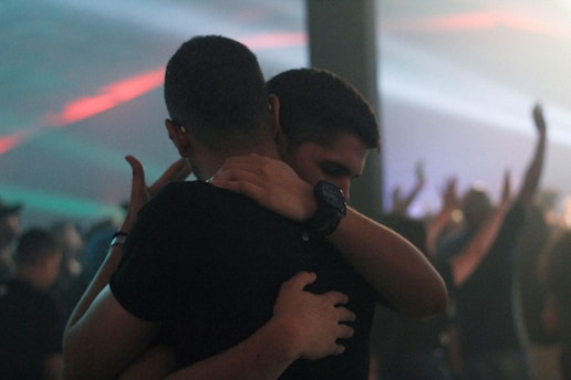 Two people are embracing in a crowded, dimly lit environment with blurred figures and colorful lighting in the background. Their expressions suggest a sense of comfort or camaraderie.