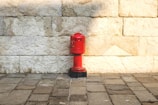 A vibrant red fire hydrant is positioned against a textured stone wall. The stone wall consists of large rectangular blocks in varying shades of beige and white. The base of the hydrant is black, and the ground is paved with gray and brown bricks.