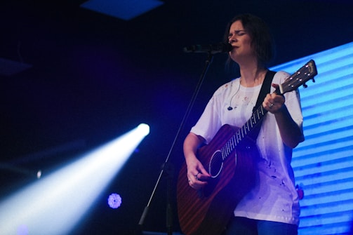 A person is playing an acoustic guitar and singing into a microphone on stage. They are lit by a spotlight, and are wearing a white shirt. The background features a blue and purple lighting, adding an atmospheric effect to the scene.