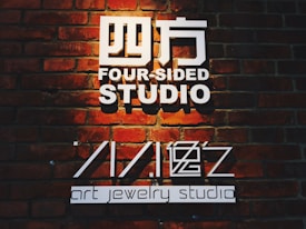The image features a sign on a brick wall that reads 'Four-Sided Studio' in large white text with additional text 'art jewelry studio'. The lighting casts a warm glow over the bricks, highlighting their rustic texture.