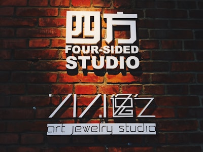 The image features a sign on a brick wall that reads 'Four-Sided Studio' in large white text with additional text 'art jewelry studio'. The lighting casts a warm glow over the bricks, highlighting their rustic texture.