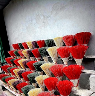 A display of colorful incense sticks arranged in several rows against a plain gray wall. The bundles are tied together and exhibit a variety of vibrant colors, including red, yellow, green, and purple. They are placed on what appears to be a series of wooden shelves or steps.