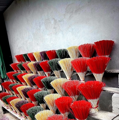 A display of colorful incense sticks arranged in several rows against a plain gray wall. The bundles are tied together and exhibit a variety of vibrant colors, including red, yellow, green, and purple. They are placed on what appears to be a series of wooden shelves or steps.