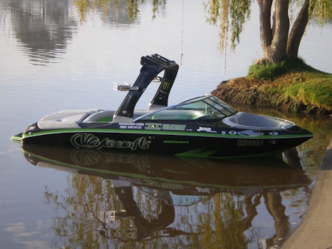 A sleek black and green speedboat is floating on calm water near the shore. The boat has various decals and branding on its sides. The shoreline has a small grassy area with a tree hanging over the water. The water reflects the surroundings, creating a mirrored image of the boat and the tree.
