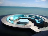 A circular outdoor seating arrangement with turquoise cushions and pillows, arranged around a matching striped table. The structure is set on a wooden deck that extends over a body of water, with views of a calm, expansive sea under a clear sky.