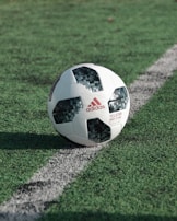 A soccer ball with a pixelated black and white pattern rests on a green grass field, partially placed on a white boundary line. The ball features the Adidas logo and branding details.