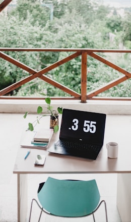 A neatly arranged workspace with a laptop displaying the time 3:55 PM, positioned on a white desk. Next to the laptop, there is a small potted plant with green leaves, several books, a pencil, a smartphone, and a white mug. The scene is set against a background featuring a wooden railing and lush greenery outside, creating a serene atmosphere.