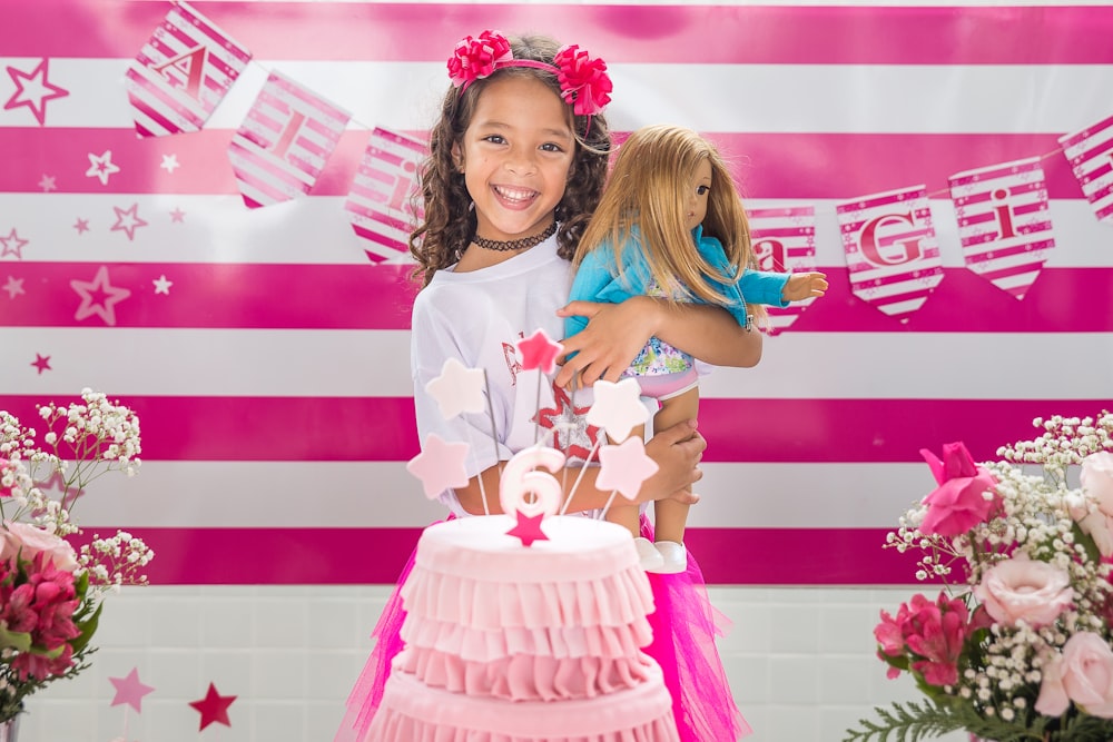 girl happily smiling holding doll with pink cake in front of her