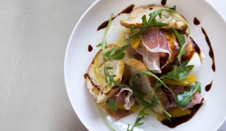 A gourmet salad is arranged on a white plate, featuring slices of prosciutto, fresh arugula leaves, toasted bread, and drizzles of balsamic reduction. There's a wedge of soft cheese and slices of citrus, possibly orange, adding a vibrant touch to the dish.