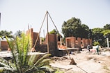 An outdoor construction site with partially built brick walls and scattered building materials. The scene includes workers engaged in construction activities under a clear sky. A large brick structure is taking shape amidst trees and greenery, hinting at a rural or suburban setting.